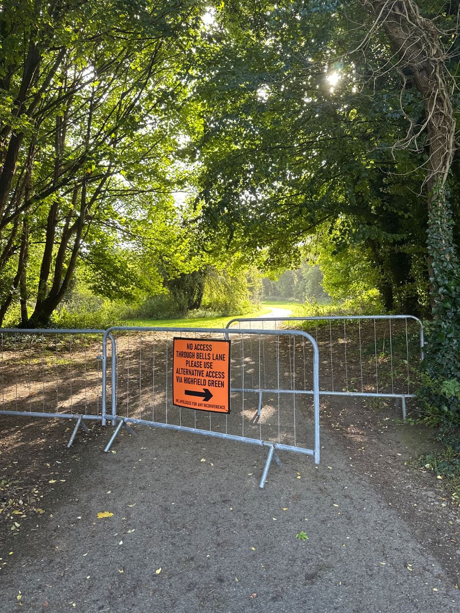 A gravel path through a wooded area blocked by metal crowd control barriers with an orange sign reading "NO ACCESS THROUGH BELLS LANE PLEASE USE ALTERNATIVE ACCESS VIA HIGHFIELD GREEN" and "NO ACCESS FOR ANY VEHICLES".